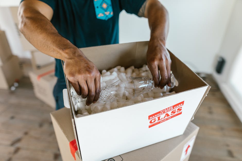 A person’s hands are shown packing a cardboard box labeled ‘Glass’ with protective white packing peanuts inside a home. The individual is placing a clear glass bottle into the box, which is situated o