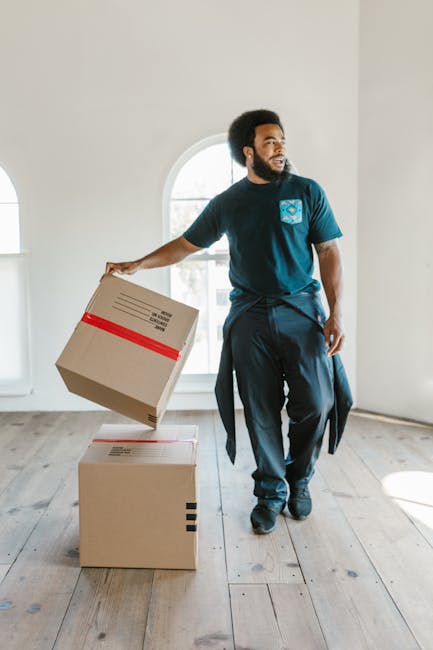 A person’s hands are shown packing a cardboard box labeled ‘Glass’ with protective white packing peanuts inside a home. The individual is placing a clear glass bottle into the box, which is situated o